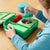Child interacting with a green lunch box containing strawberries and other food items on a wooden table.