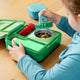 Child interacting with a green lunch box containing strawberries and other food items on a wooden table.