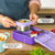 Purple lunch box with compartments and a thermos for hot food, on a wooden table.