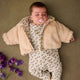 Baby lying on a brown surface wearing a floral outfit and beige coat with purple flowers nearby.