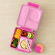 Pink bento box with compartments filled with fruits and vegetables on a beige surface